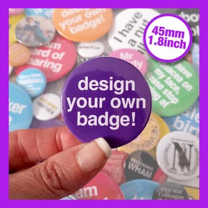 Op de afbeelding: Een hand houdt een paarse knop van 4,5 cm met witte tekst die "design your own badge!" luidt. De knop is omringd door andere knoppen met verschillende ontwerpen en tekst.