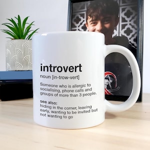 Op de afbeelding: Witte keramische mok met een zwart-witte definitie van het woord "introvert" erop gedrukt. De definitie omvat de uitspraak, een beschrijving van een introvert en een lijst met veelvoorkomende introvert gedrag.