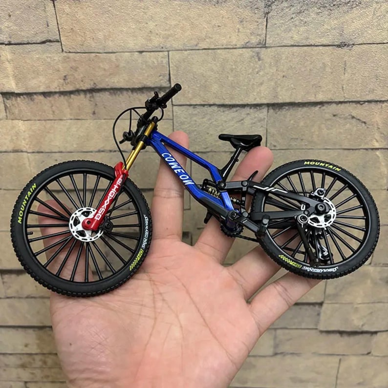 May include: A miniature mountain bike with a blue frame, black tires, and red accents. The bike has the text "COME ON" on the frame and "MOUNTAIN" on the tires. The bike is held in a person's hand.