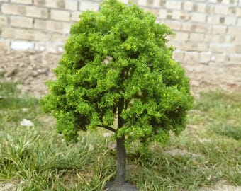 May include: A miniature model tree with vibrant green foliage and a dark brown trunk. The tree stands in a grassy area, with a brick wall in the background. This small tree is ideal for model train sets or miniature landscapes.