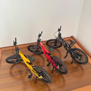 Könnte beinhalten: Vier Miniatur-Mountainbikes in Gelb, Rot und Schwarz werden auf einer Holzoberfläche ausgestellt. Jedes Fahrrad hat schwarze Reifen, einen schwarzen Sitz und einen Ständer. Die Fahrräder sind in verschiedenen Winkeln angeordnet.