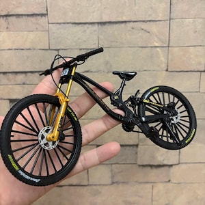 Könnte beinhalten: Ein Miniatur-Mountainbike in Schwarz mit goldenen Federgabeln und schwarzen Reifen. Das Fahrrad hat die Nummer 33 auf der Vorderseite und das Wort "MOUNTAIN" ist auf den Reifen zu sehen. Das Fahrrad wird in einer Hand gehalten.