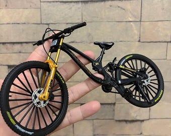 May include: A miniature black mountain bike with gold suspension forks and black tires. The bike has the number 33 on the front and the word "MOUNTAIN" is visible on the tires. The bike is held in a person's hand.
