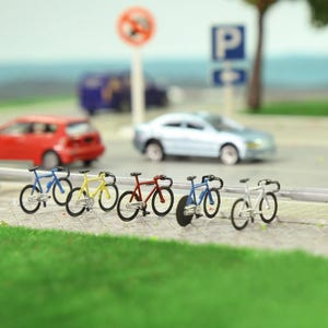 以下が含まれることがあります： 青、黄、赤、白のミニチュア自転車が歩道に並んでいます。小さな自転車は黒いタイヤとハンドルバーが付いています。背景には赤い車、青いバン、シルバーの車があります。