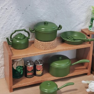 May include: A miniature kitchen set with olive green cookware, including a kettle, pots, and a frying pan with a spatula. The set is displayed on a wooden shelf alongside miniature canned goods and a jar of pickles.