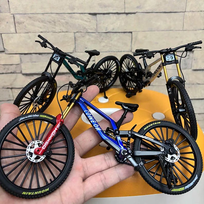 May include: Miniature mountain bikes in blue, green, and gold. The blue bike has the text "COME ON" on the frame and "MOUNTAIN" on the tire. The bikes have black tires and frames, with gold and red accents. Each bike has a number on the front.