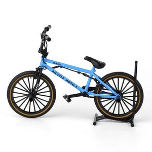 Op de afbeelding: Een lichtblauwe BMX-fiets met zwarte banden en een zwart zadel. De woorden "RACE" en "BICYCLE MOTO-X" staan op het frame gedrukt. De fiets wordt op een zwarte standaard gepresenteerd.