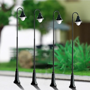 Könnte beinhalten: Vier Miniatur-Straßenlaternen in Schwarz mit weißen Kugellampen. Die Lampen haben ein klassisches Design mit gebogenem Oberteil und Sockel. Der Hintergrund ist unscharf, was auf eine Außenumgebung hindeutet, möglicherweise eine Modelleisenbahn oder ein Puppenhaus.