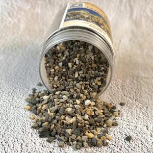 以下が含まれることがあります： 透明なプラスチックの瓶が傾いており、小さなマルチカラーの砂利をテクスチャ加工された表面にこぼしています。砂利には、グレー、ブラウン、ベージュの色合いが含まれています。瓶にはテキストと風景画像が記載されたラベルが付いています。