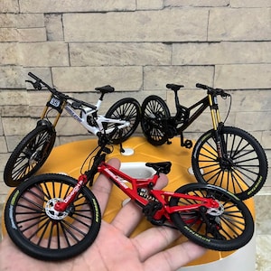 Könnte beinhalten: Miniatur-Mountainbikes in Rot, Schwarz und Weiß, mit schwarzen Reifen und aufwendigen Details. Die Fahrräder werden auf einer gelben Oberfläche präsentiert, um ihr Design und ihre Eigenschaften hervorzuheben. Auf den Fahrrädern steht das Wort "Simulation".