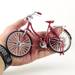 May include: A miniature burgundy bicycle with silver accents. The bike features a classic design with a luggage rack, fenders, and a comfortable seat. The wheels have silver spokes and black tires. The handlebars and seat are burgundy.