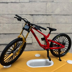 K&ouml;nnte beinhalten: Ein Miniatur-Mountainbike in Rot und Gelb mit schwarzen Reifen und Lenker. Das Fahrrad hat die Nummer 41 auf der Vorderseite und das Wort "FORK" auf der Vorderradaufh&auml;ngung. Das Fahrrad steht auf einer gelben Oberfl&auml;che.