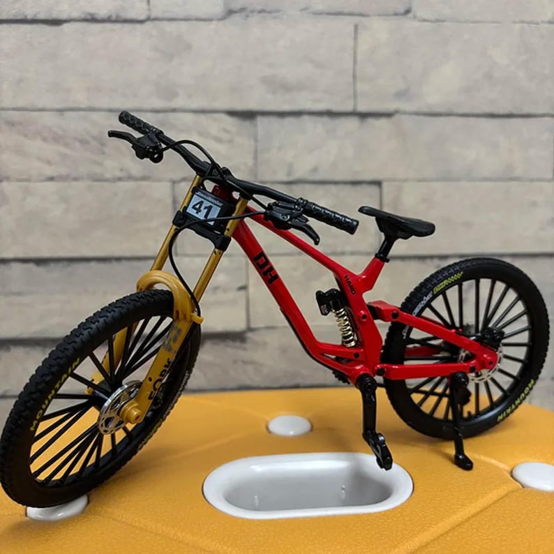 K&ouml;nnte beinhalten: Ein Miniatur-Mountainbike in Rot und Gelb mit schwarzen Reifen und Lenker. Das Fahrrad hat die Nummer 41 auf der Vorderseite und das Wort "FORK" auf der Vorderradaufh&auml;ngung. Das Fahrrad steht auf einer gelben Oberfl&auml;che.