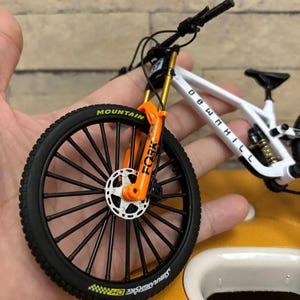 Puede incluir: Una bicicleta de monta&ntilde;a en miniatura blanca y negra con horquillas de suspensi&oacute;n naranjas y neum&aacute;ticos negros. El neum&aacute;tico delantero tiene la palabra "MOUNTAIN". El cuadro tiene las palabras "DOWNHILL".
