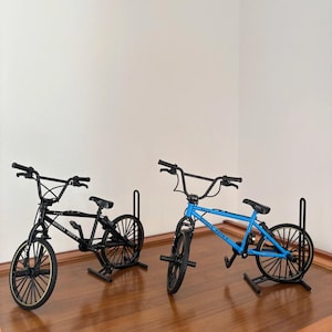 Op de afbeelding: Twee miniatuur BMX-fietsen, &eacute;&eacute;n zwart en &eacute;&eacute;n blauw, worden tentoongesteld op een houten oppervlak. De zwarte fiets heeft goudkleurige velgen, terwijl de blauwe fiets zwarte velgen heeft. Beide fietsen hebben zwarte sturen en zadels. De tekst "Bicycle Moto X" is zichtbaar.
