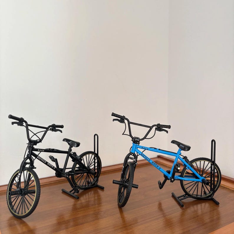 Op de afbeelding: Twee miniatuur BMX-fietsen, &eacute;&eacute;n zwart en &eacute;&eacute;n blauw, worden tentoongesteld op een houten oppervlak. De zwarte fiets heeft goudkleurige velgen, terwijl de blauwe fiets zwarte velgen heeft. Beide fietsen hebben zwarte sturen en zadels. De tekst "Bicycle Moto X" is zichtbaar.