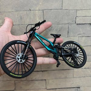 May include: A miniature mountain bike in teal and black, held in a hand. The bike features black tires with "MOUNTAIN" text, a black frame, and a teal-colored body. The wheels have multiple spokes.