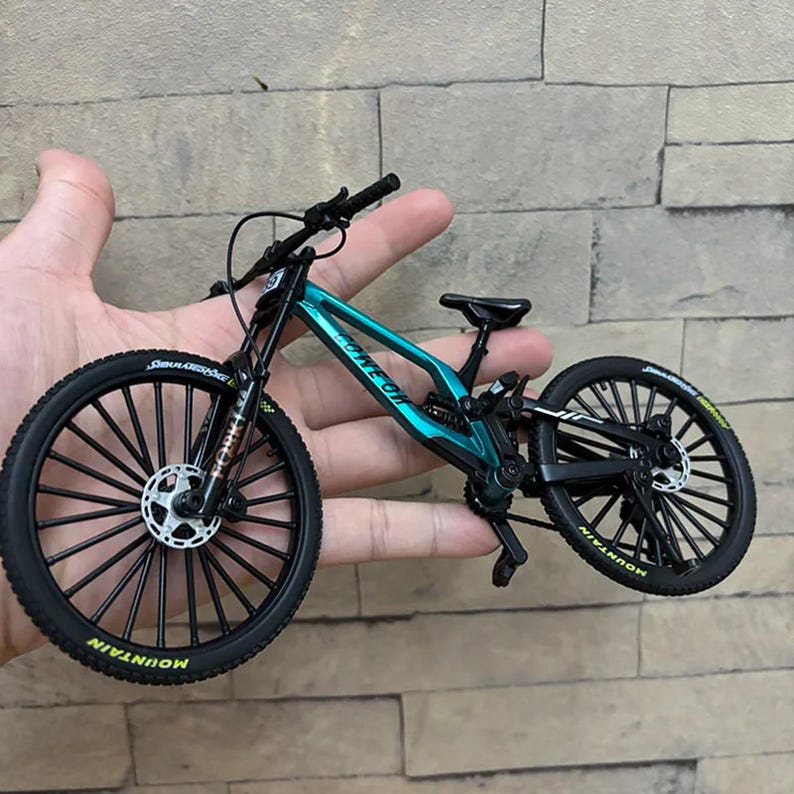 May include: A miniature mountain bike in teal and black, held in a hand. The bike features black tires with "MOUNTAIN" text, a black frame, and a teal-colored body. The wheels have multiple spokes.