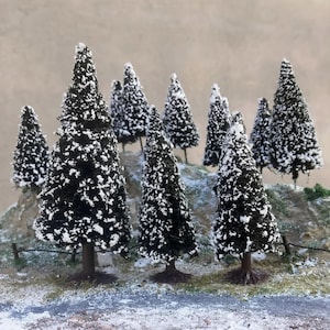 May include: A model winter forest scene featuring dark green trees covered in white snow. The miniature trees are set on a small hill with a snowy ground, a small fence, and a road, against a light brown backdrop.