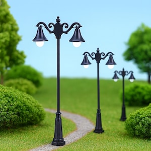 May include: Miniature black street lamps with two bell-shaped lights, set in a garden scene with a winding path and green foliage. The lamps have ornate detailing and are evenly spaced.