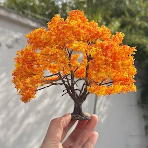May include: A miniature tree with vibrant orange and yellow foliage, resembling autumn colours. The tree has a dark brown trunk and branches, and is held in a hand. The tree is a decorative item.