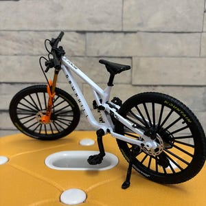Puede incluir: Una bicicleta de monta&ntilde;a blanca en miniatura con neum&aacute;ticos negros y un asiento negro. La horquilla delantera es naranja y las ruedas tienen radios negros. La bicicleta tiene las palabras "Downhill" y "Mountain".