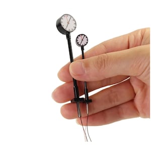 Puede incluir: Dos relojes en miniatura en blanco y negro con tallos y cables negros. Los relojes tienen esferas blancas con números y manecillas negras. Los relojes se sostienen en una mano sobre un fondo blanco.