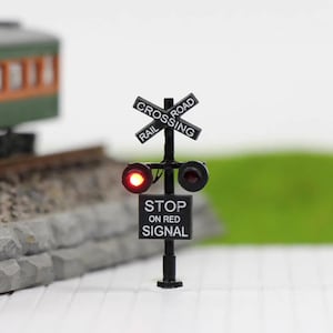 Puede incluir: Una señal de cruce ferroviario negra en miniatura con una luz roja encendida. El letrero dice "RAIL CROSSING" y "STOP ON RED SIGNAL". La señal se encuentra sobre un fondo borroso con un tren modelo.