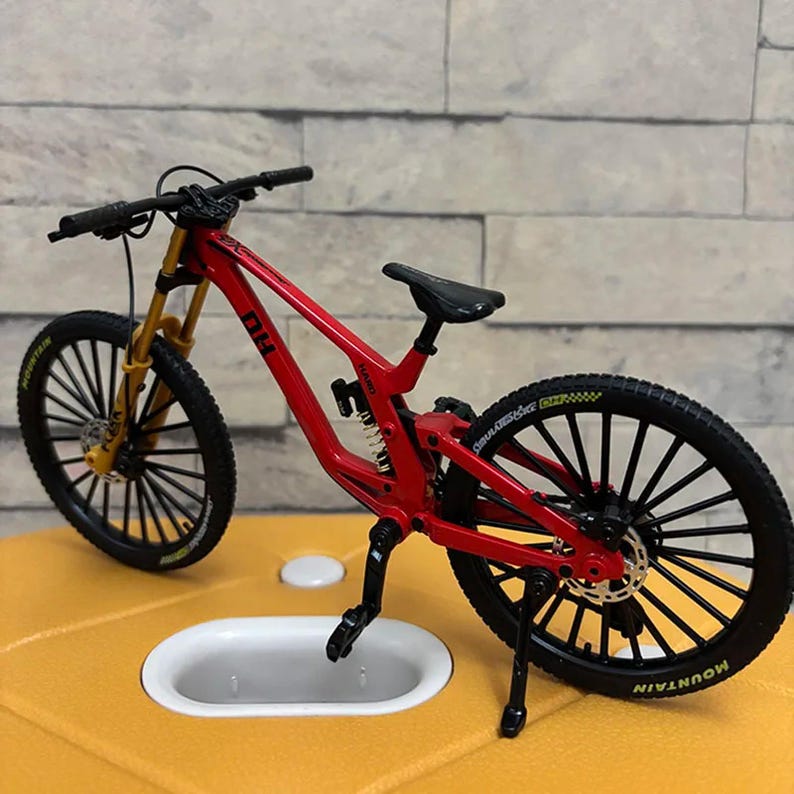 K&ouml;nnte beinhalten: Ein Miniatur-Mountainbike in Rot mit schwarzen Reifen und goldenen Federgabeln. Das Fahrrad hat einen schwarzen Sattel und Lenker. Auf den R&auml;dern steht das Wort "Mountain" auf den Reifen. Das Fahrrad ist ein detailliertes Modell.