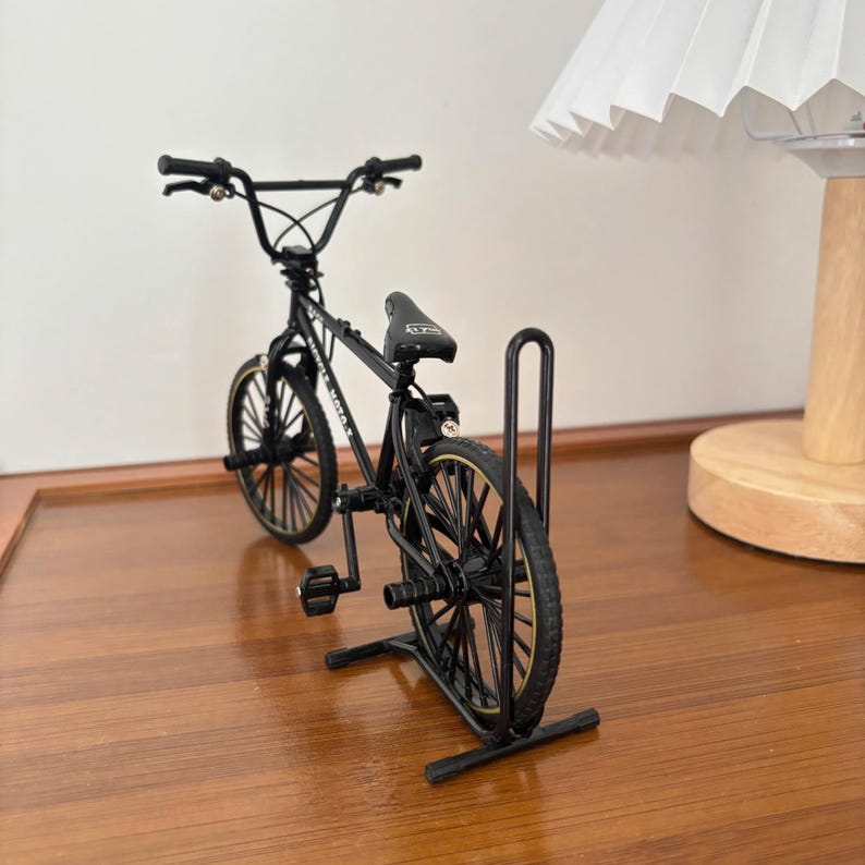 Op de afbeelding: Een miniatuur zwarte BMX-fiets met goudkleurige accenten op de banden en een zwart zadel. De fiets wordt tentoongesteld op een zwarte metalen standaard. Het stuur is zwart en de wielen hebben meerdere spaken.
