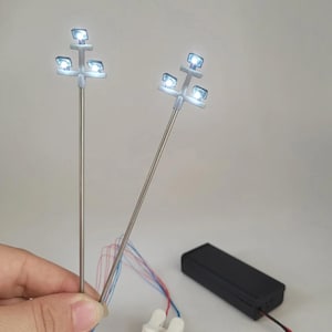 May include: Two miniature street lamps with bright white LED lights. Each lamp has three lights on a white fixture atop a silver pole. Wires of various colours extend from the base of the lamps. A black battery pack and a white connector are also visible.