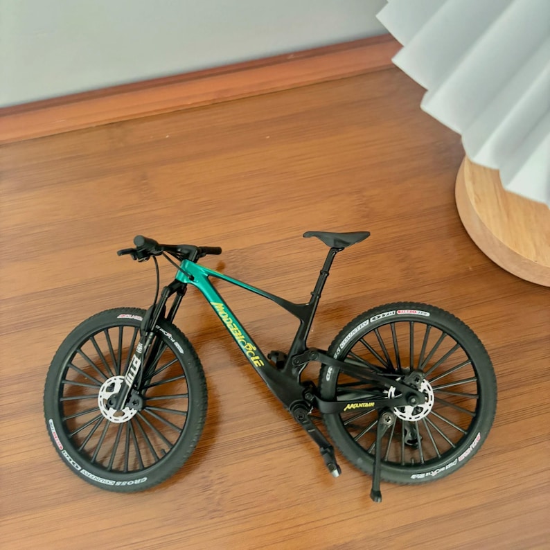 May include: A miniature mountain bike with a black frame and teal accents. The bike has black tires, a black seat, and the words "modaric cycle" are printed on the frame. The bike is on a wooden surface.