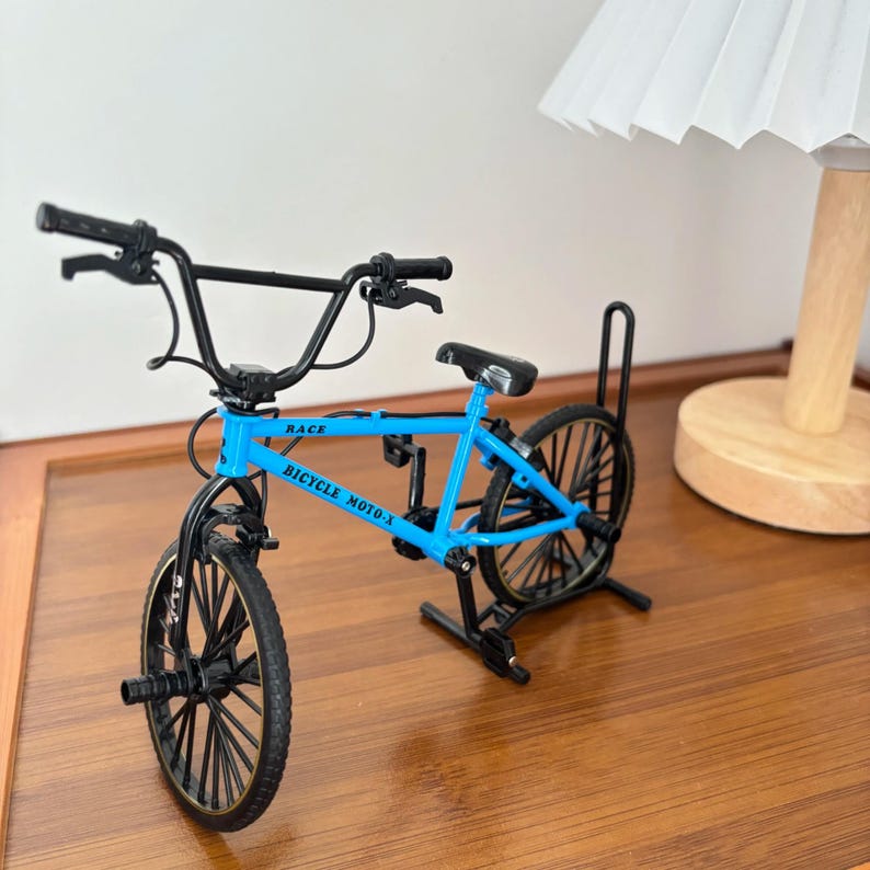 Op de afbeelding: Een miniatuur BMX-fiets in helderblauw, met zwarte stuur, wielen en zadel. Het frame toont de tekst "RACE BICYCLE MOTO-X". De fiets is ontworpen met ingewikkelde details, wat een realistische uitstraling geeft.