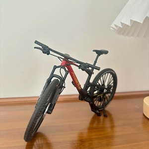 May include: A miniature mountain bike with a red and black frame. The bike has black tires, a black seat, and a black handlebar. The text "POWERBICYCLE" is printed on the frame. The bike is displayed on a wooden surface.