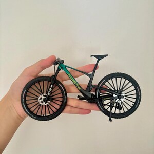 May include: A miniature mountain bike with a green and black frame, black tires, and a black seat. The bike has a detailed design, including disc brakes and a suspension fork. The wheels have multiple spokes.