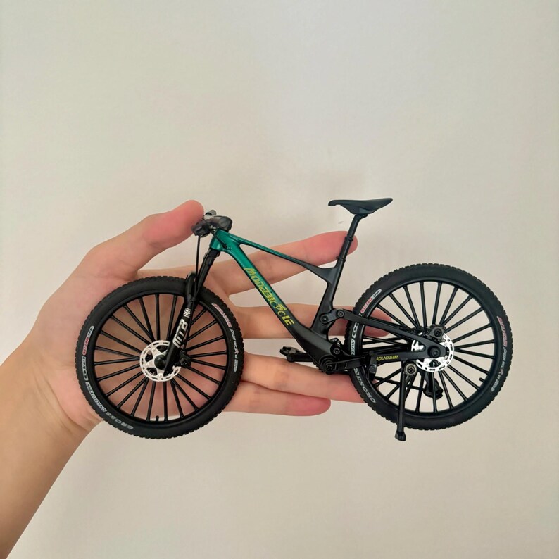 May include: A miniature mountain bike with a green and black frame, black tires, and a black seat. The bike has a detailed design, including disc brakes and a suspension fork. The wheels have multiple spokes.