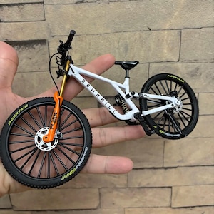 Puede incluir: Una bicicleta de monta&ntilde;a en miniatura blanca y negra con horquillas de suspensi&oacute;n naranjas y doradas. Los neum&aacute;ticos son negros con la palabra "MOUNTAIN" en amarillo. El cuadro tiene la palabra "DOWNHILL" impresa.