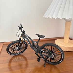 Könnte beinhalten: Ein Miniatur-Mountainbike in Schwarz mit goldenen Federgabeln und schwarzen Reifen. Das Fahrrad hat einen kleinen schwarzen Sattel und einen Ständer. Die Räder haben schwarze Speichen und das Fahrrad steht auf einer Holzoberfläche.