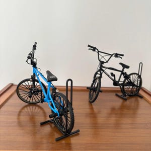 Op de afbeelding: Twee miniatuur BMX-fietsen, &eacute;&eacute;n blauw en &eacute;&eacute;n zwart, worden gepresenteerd op een houten oppervlak. De blauwe fiets is naar de kijker gekanteld, waarbij de details worden benadrukt. De zwarte fiets staat erachter. Beide fietsen hebben zwarte banden en zadels.