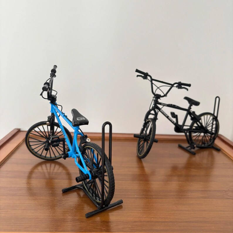 Op de afbeelding: Twee miniatuur BMX-fietsen, &eacute;&eacute;n blauw en &eacute;&eacute;n zwart, worden gepresenteerd op een houten oppervlak. De blauwe fiets is naar de kijker gekanteld, waarbij de details worden benadrukt. De zwarte fiets staat erachter. Beide fietsen hebben zwarte banden en zadels.