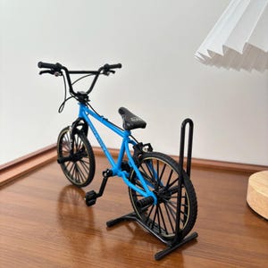 Op de afbeelding: Een miniatuur BMX-fiets in helderblauw, met zwarte banden en een zwart stuur. De fiets wordt tentoongesteld op een zwarte standaard, die het gedetailleerde ontwerp benadrukt. De fiets heeft een zwart zadel en pedalen.