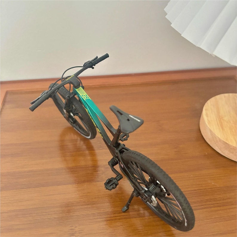 May include: A miniature mountain bike with a black frame and tires. The bike features a teal and green accent on the frame and a black seat. The handlebars are black, and the wheels have silver spokes. The bike is resting on a wooden surface.
