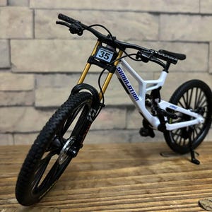 Könnte beinhalten: Ein weißes Miniatur-Mountainbike mit schwarzen Reifen und Lenker. Das Fahrrad hat goldene Federgabeln und das Wort "SIMULATION" auf dem Rahmen aufgedruckt. Eine kleine Zahl "35" ist vorne sichtbar.