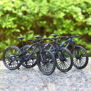 Könnte beinhalten: Vier Miniatur-Mountainbikes in Schwarz, Braun und Blau werden gezeigt. Jedes Fahrrad hat schwarze Reifen, einen schwarzen Rahmen und einen schwarzen Sattel. Die Fahrräder sind in einer Reihe angeordnet, wobei die Vorderräder zum Betrachter zeigen. Der Hintergrund ist ein verschwommenes Grün.