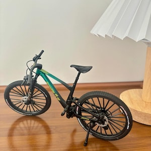 May include: A miniature mountain bike with a green and black frame. The bike has black tires, a black seat, and a kickstand. The wheels have white lettering. The bike is a detailed model.