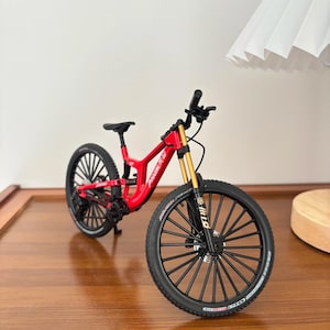 Puede incluir: Una bicicleta de montaña roja en miniatura con neumáticos negros y horquillas de suspensión doradas. La bicicleta tiene un asiento y manillar negros. Los neumáticos tienen letras blancas. La bicicleta está sobre una superficie de madera.
