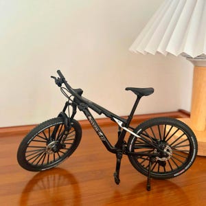 May include: A black mountain bike model with a detailed design. The bike features black tires, a black frame, and a black seat. The wheels have multiple spokes. The bike is displayed on a wooden surface.