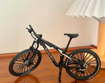 May include: A black mountain bike model with a detailed design. The bike features black tires, a black frame, and a black seat. The wheels have multiple spokes. The bike is displayed on a wooden surface.