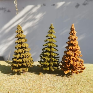 May include: Three model pine trees in shades of green and brown, set on a bed of tan-coloured material. The trees have a detailed, textured appearance, with a neutral background. These miniature trees are suitable for model railway layouts or dioramas.