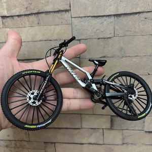 May include: A miniature mountain bike with a silver frame, black tires, and gold suspension. The tires have the word "MOUNTAIN" in yellow. The bike is held in a hand against a brick wall background.